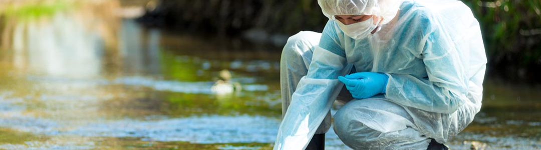a person wearing a hazmat suit collects a water sample from a stream